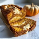 A loaf of pumpkin bread with cream cheese filling, sliced on a wooden board. Decorative pumpkin in the background.