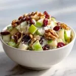 A white bowl filled with crisp apple salad, featuring green apples, walnuts, and cranberries.