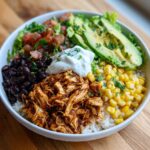 Overhead view of a vibrant chicken burrito bowl with rice, chicken, beans, corn, avocado, salsa, and sour cream.