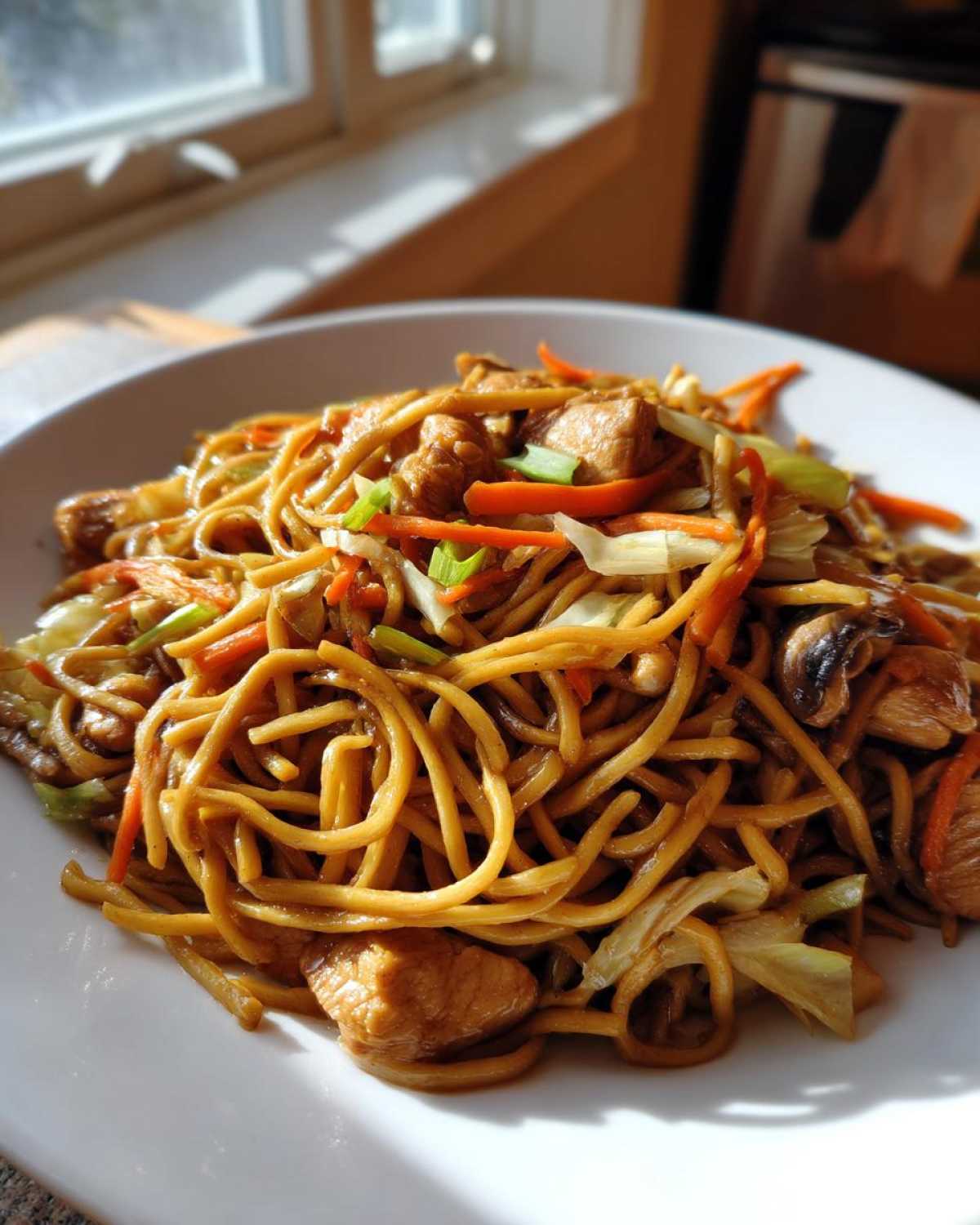 A close-up of a serving of homemade chicken chow mein recipe with noodles, chicken chunks, carrots, and cabbage.