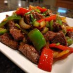 A close-up of glossy, saucy pepper steak strips mixed with bright red and green bell peppers and onions.