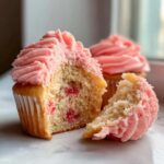 Close-up of a strawberry cupcake cut in half showing the moist interior with strawberry pieces and pink frosting.