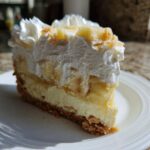 A tall slice of banana cream cheesecake with a graham cracker crust, creamy filling, banana slices, and whipped cream topping.