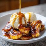 Caramel sauce being poured over caramelized bananas and vanilla ice cream in a white bowl, classic Bananas Foster dessert.