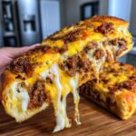 A hand pulling apart a slice of cheesy garbage bread, showing melted mozzarella and cheddar dripping over a savory meat filling.