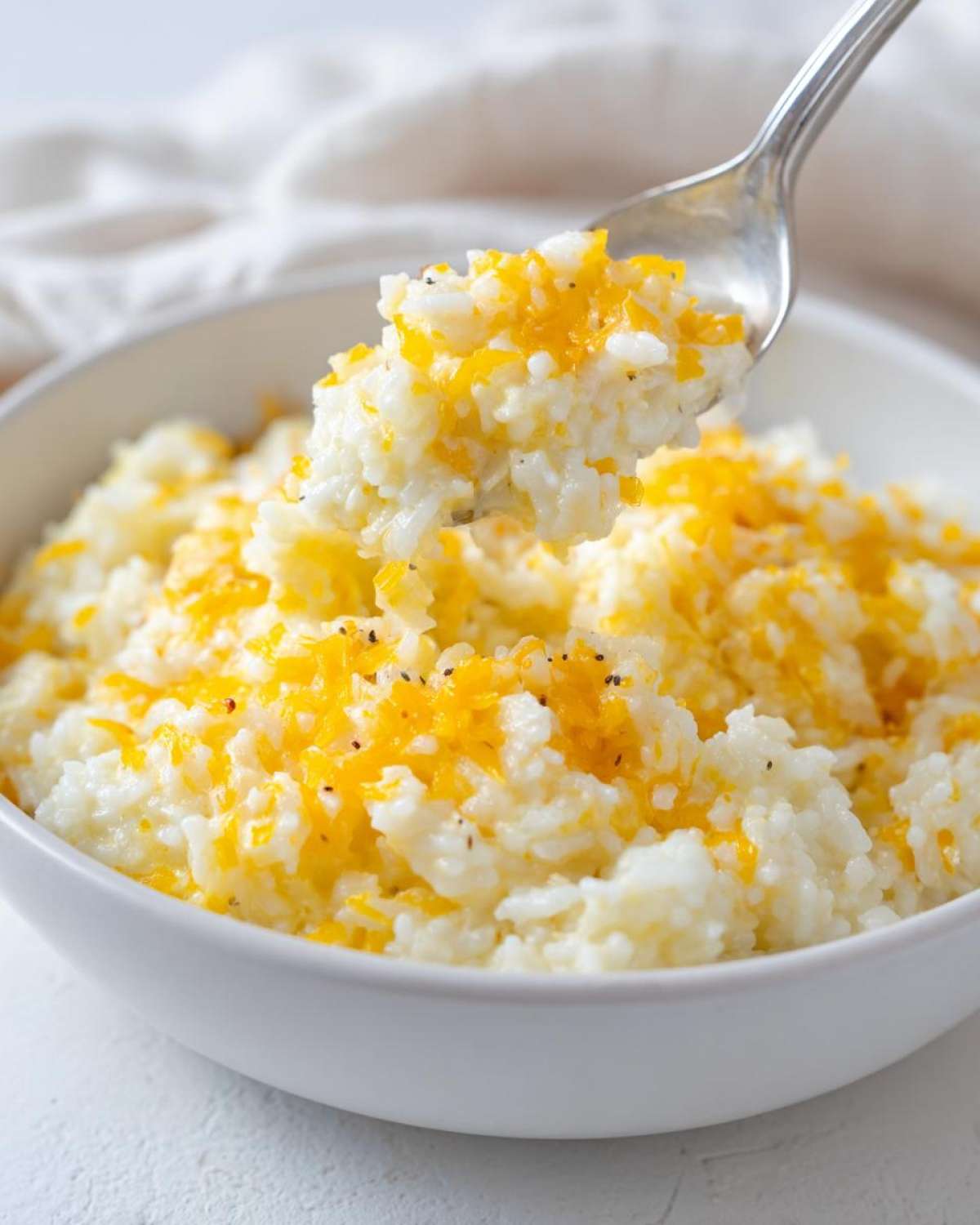 A spoonful of creamy, white cheesy rice topped with melted shredded cheddar cheese being lifted from a white bowl.