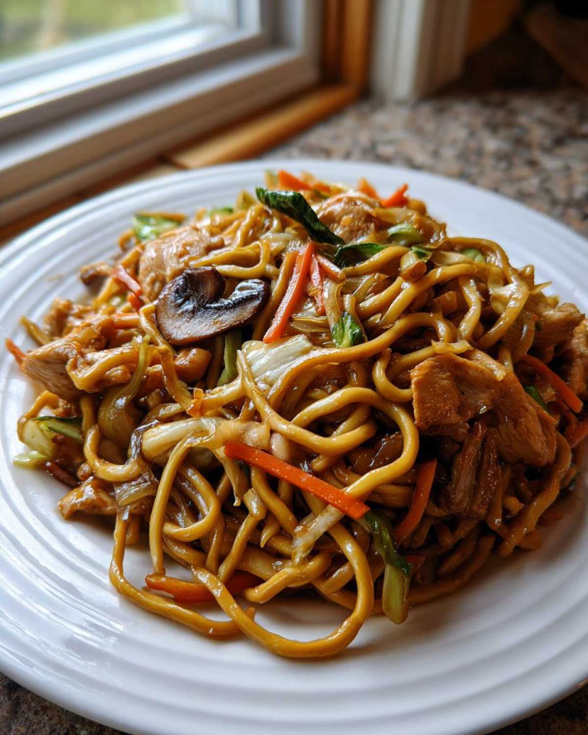 A close-up of a generous serving of homemade chicken chow mein recipe with thick noodles, chicken pieces, carrots, and mushrooms.
