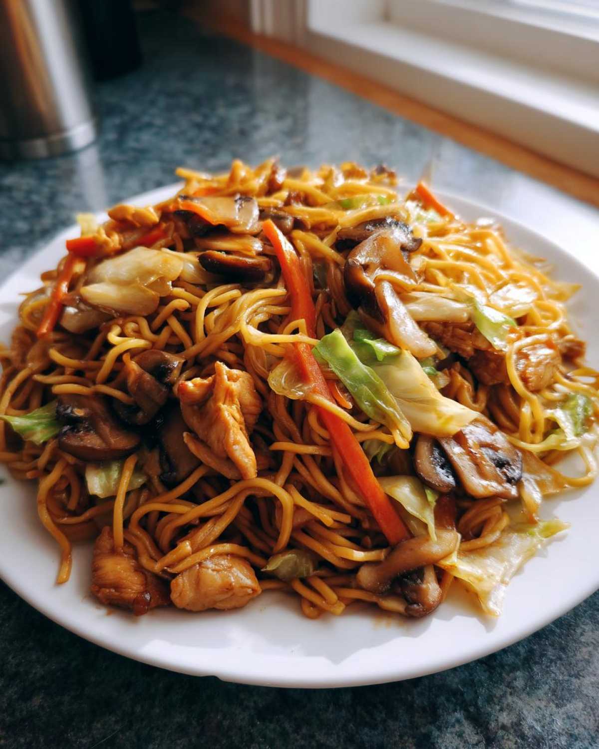 A close-up of a white plate filled with homemade chicken chow mein recipe, featuring noodles, chicken pieces, mushrooms, and cabbage.