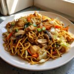 A close-up of a generous serving of chicken chow mein recipe, featuring noodles, chicken pieces, mushrooms, and cabbage.
