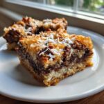A close-up of two squares of rich magic cookie bars topped with toasted coconut and chocolate chips on a white plate.
