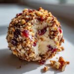 Close-up of a creamy cranberry pecan cheese ball coated in chopped nuts, with a small portion removed.