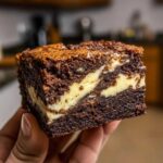A close-up of a rich, fudgy square of cream cheese brownies with a visible swirl of cheesecake filling.