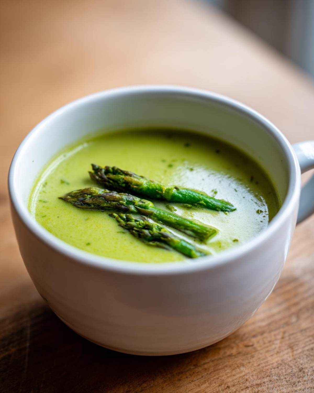 Close-up of a white mug filled with bright green asparagus soup, topped with three cooked asparagus spears.