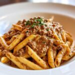A close-up of a white bowl filled with creamy beef pasta, featuring penne noodles coated in a rich sauce.