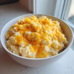 A white bowl filled with creamy cheesy rice topped with melted orange cheddar cheese and black pepper.