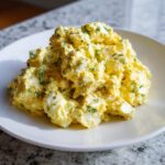 A mound of creamy, yellow egg salad mixed with chopped green herbs served on a white plate.