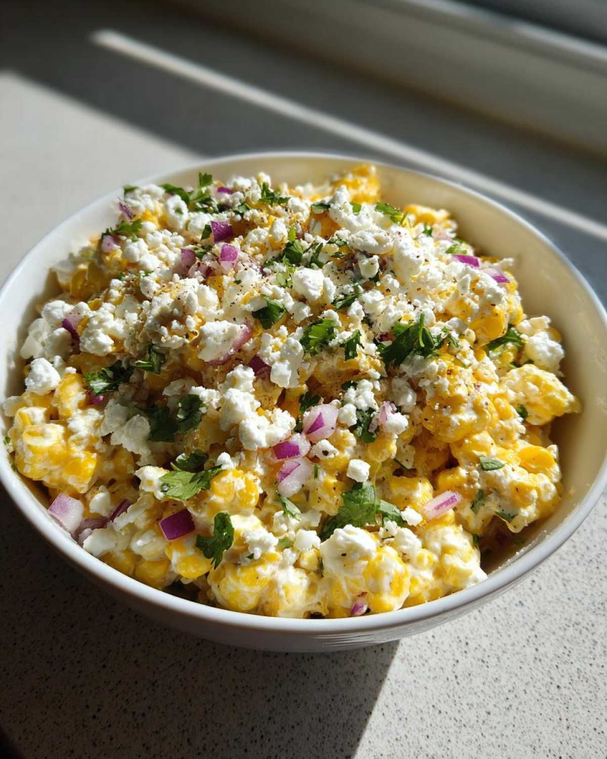 Close-up of a white bowl filled with creamy street corn salad, topped with crumbled white cheese, red onion, and cilantro.