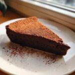 A perfect slice of rich, dark flourless chocolate torte dusted heavily with cocoa powder on a white plate.