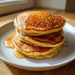 A stack of three fluffy coconut flour pancakes drizzled generously with golden syrup on a white plate.