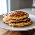 A close-up of a stack of three fluffy greek yogurt pancakes drizzled with syrup on a white plate.