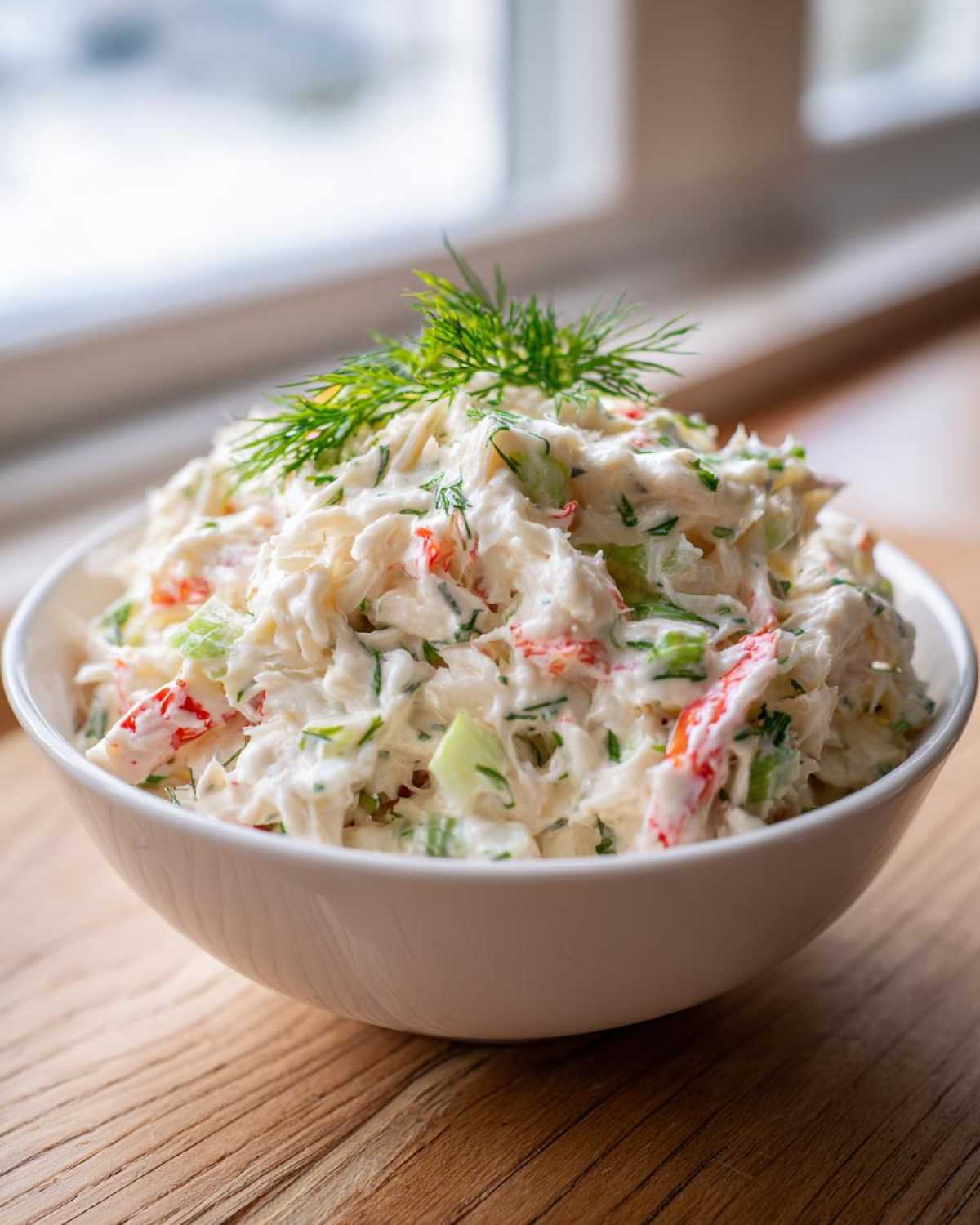A close-up of creamy crab salad mixed with celery and herbs, served in a white bowl and garnished with fresh dill.