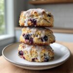 Three glazed blueberry scones stacked vertically on a light ceramic plate, showing juicy berries inside.