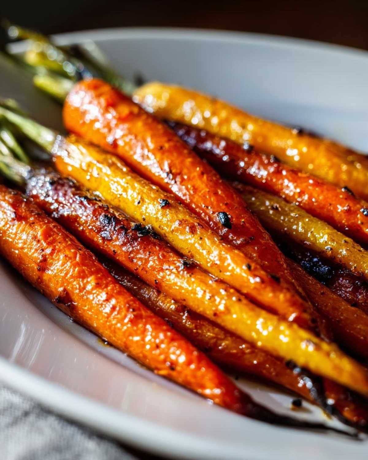 A close-up of several glazed, caramelized roasted carrots served on a light plate, showing slight charring and green tops.