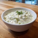 A white bowl filled with creamy homemade tartar sauce recipe, garnished with fresh dill, sitting on a wooden surface.