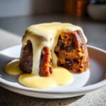 Close-up of a moist, dark plum pudding topped generously with thick, creamy custard sauce.