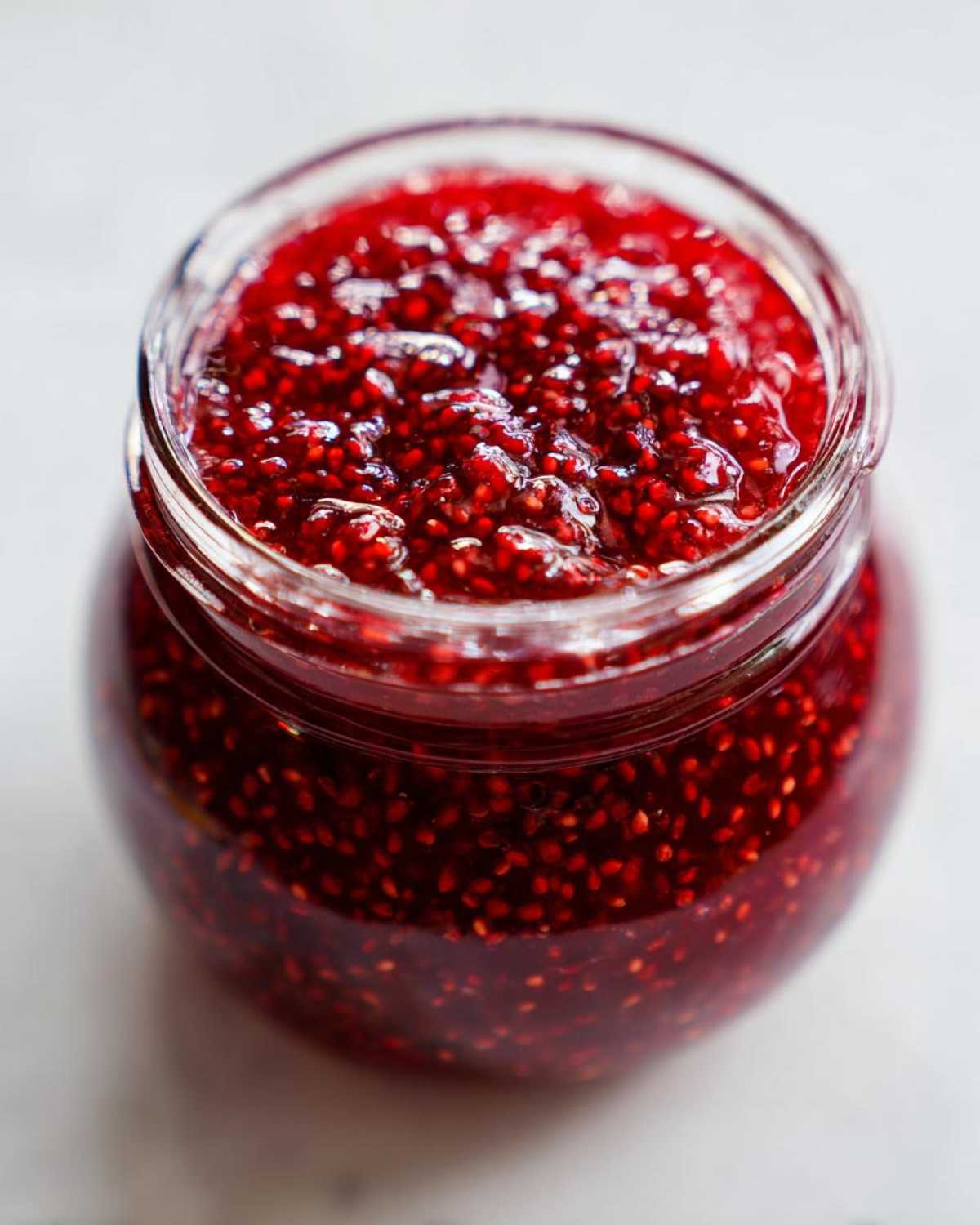 Close-up overhead view of a glass jar filled with vibrant, thick raspberry jam, showing visible seeds.
