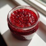Close-up overhead view of a glass jar filled with vibrant, thick raspberry jam, showing many seeds.