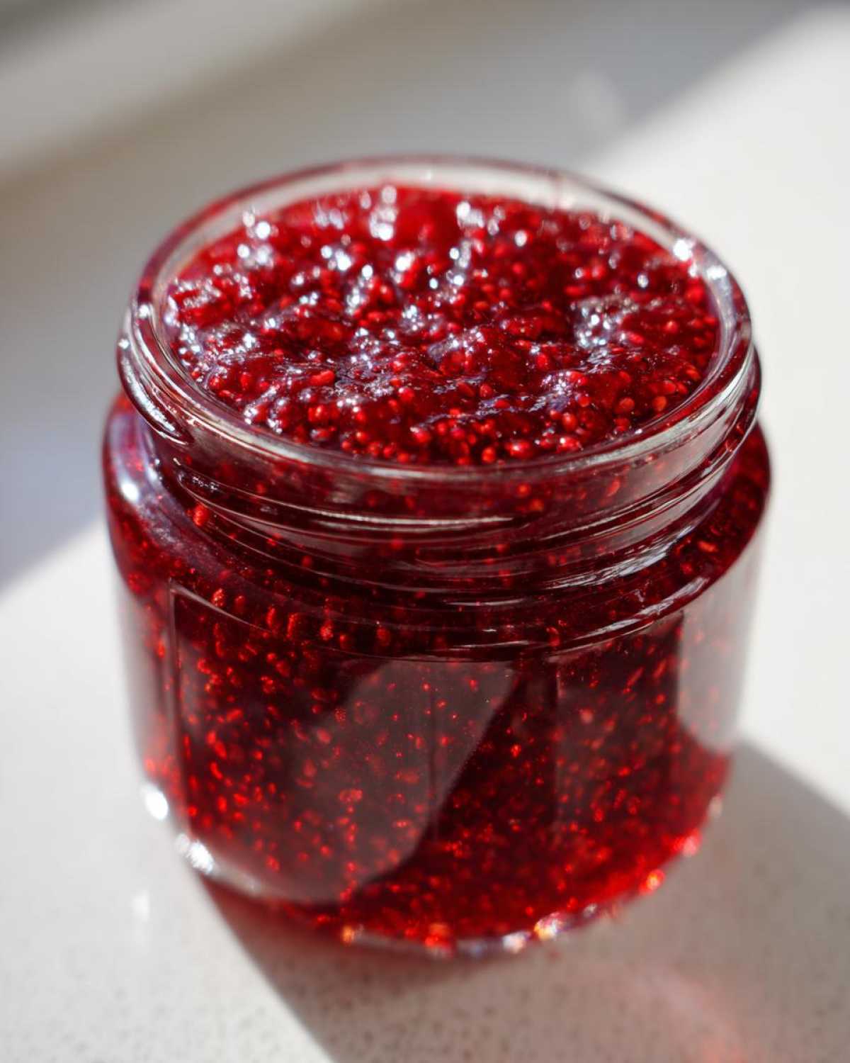 Close-up of a glass jar filled with vibrant, textured raspberry jam, glistening in the sunlight.