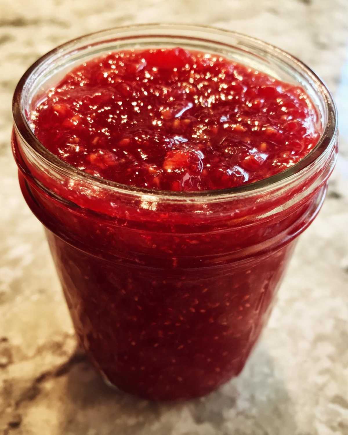 Close-up of a glass jar filled with vibrant red, textured freezer jam, likely raspberry or strawberry.