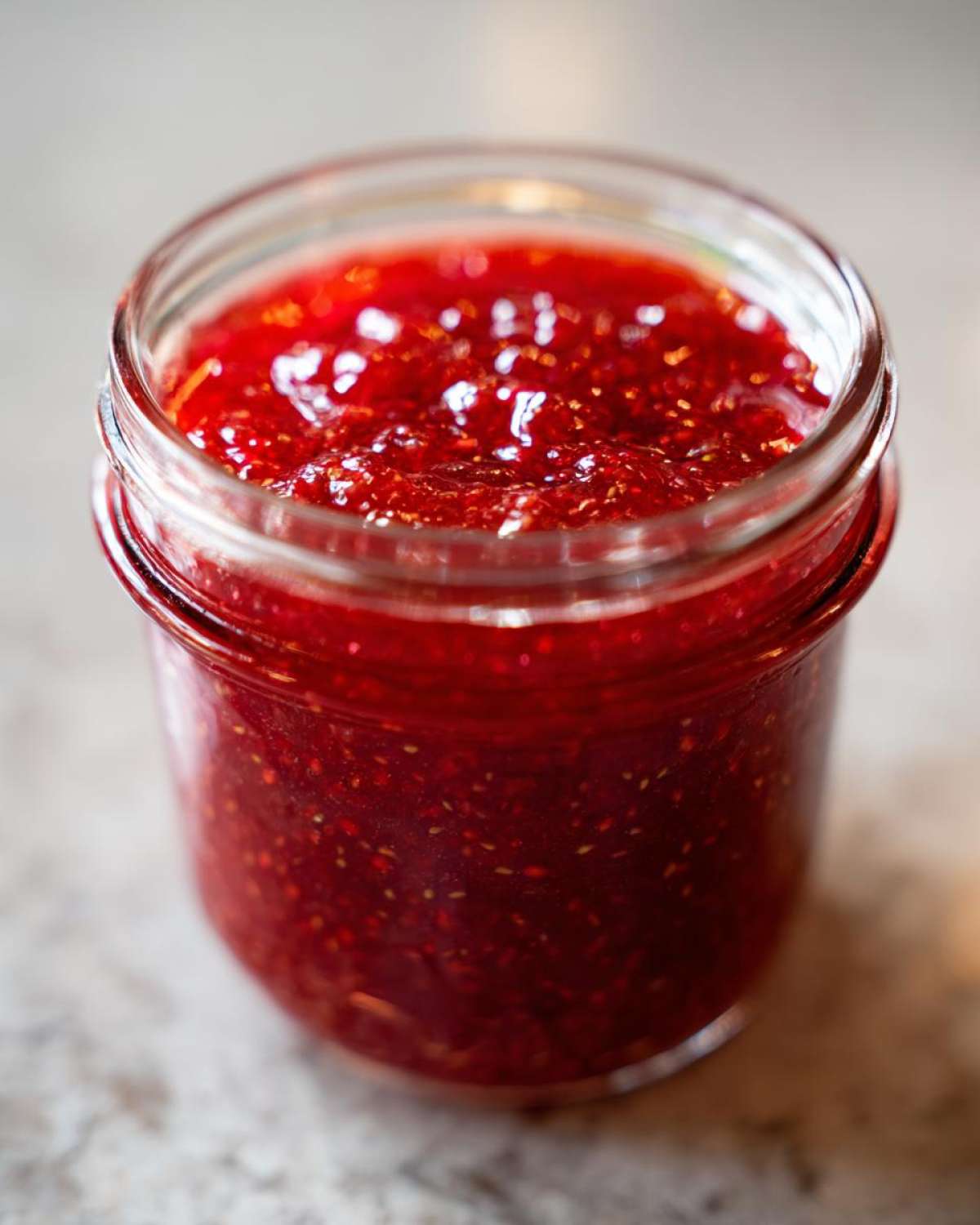 Close-up of a glass jar filled with bright red, textured freezer jam, showing visible seeds.