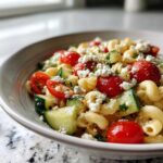 Close-up of a bowl filled with mediterranean pasta salad featuring elbow macaroni, cherry tomatoes, cucumber chunks, and crumbled feta cheese.