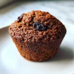 A close-up, appetizing shot of a single, dark brown, moist bran muffin topped with two raisins.
