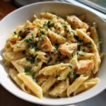 Close-up of penne pasta and chicken pieces coated in a rich garlic parmesan sauce, topped with fresh parsley.
