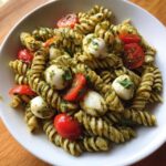 A white bowl filled with pesto pasta salad featuring rotini pasta, small mozzarella balls, and halved cherry tomatoes.