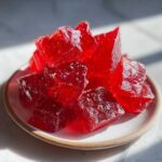 Pile of vibrant, translucent red cinnamon rock candy chunks glistening on a small white plate.