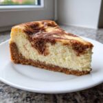 A close-up of a rich slice of snickerdoodle cheesecake featuring a thick graham cracker crust and a cinnamon-swirled top.