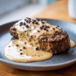A thick-cut steak generously topped with creamy peppercorn sauce and whole black peppercorns.