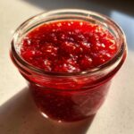 Close-up of a small glass jar filled with bright red, textured freezer jam, glistening in the sunlight.