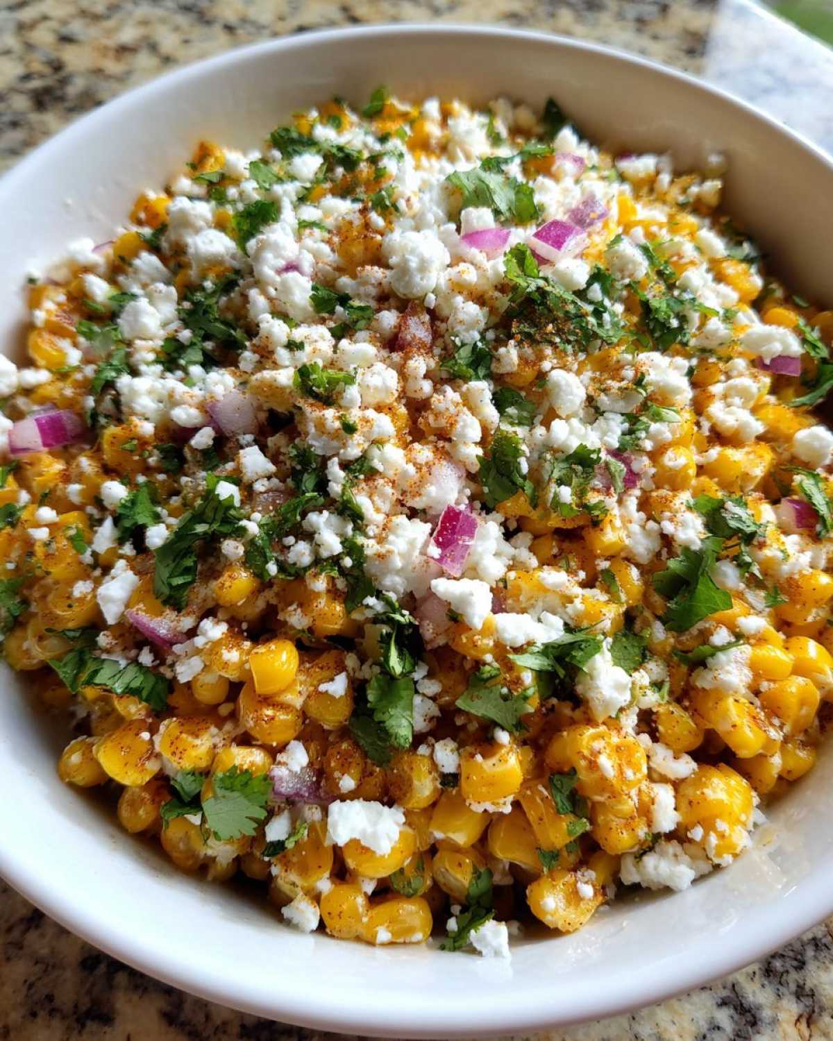 A close-up view of a vibrant street corn salad topped with crumbled white cheese, chopped cilantro, red onion, and chili powder.