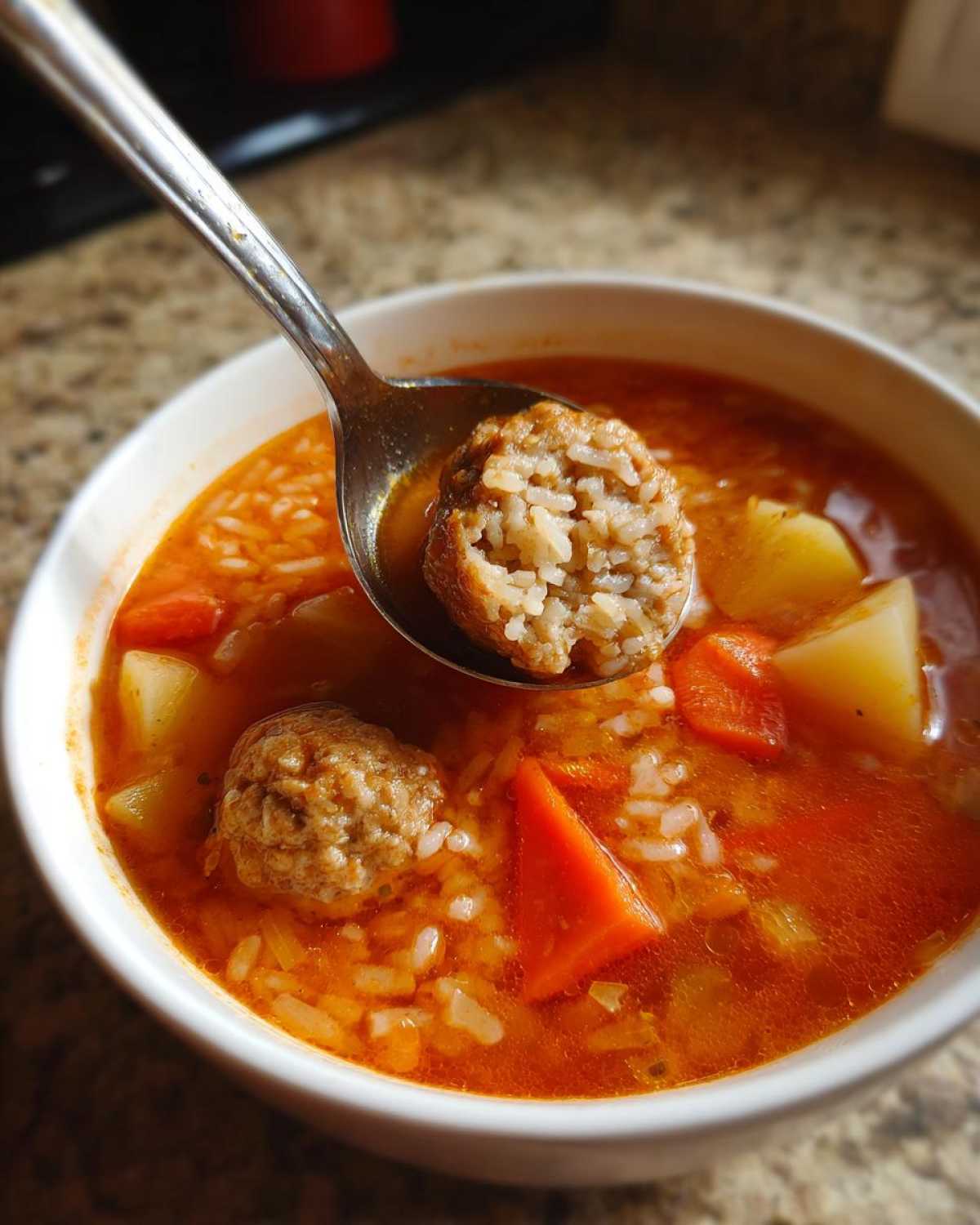 A spoonful of albondigas soup showing a cross-section of a rice-filled meatball above a bowl of rich broth with carrots and potatoes.