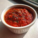 Close-up of vibrant red, chunky pizza sauce seasoned with dried herbs in a small white bowl.