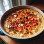 A creamy, baked street corn dip in a round dish, topped with crumbled white cheese and a dusting of red chili powder.