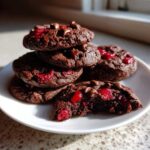 A stack of rich, dark chocolate cherry cookies topped with chocolate chips, with one cookie in front having a bite taken out.