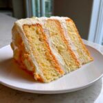 A tall slice of layered Creamsicle Cake featuring orange and yellow sponge layers with white frosting.