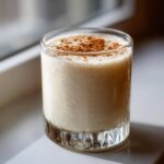 A glass of creamy, frothy coquito topped with a dusting of ground cinnamon, sitting on a bright windowsill.