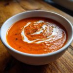 Close-up of a bowl of vibrant orange tomato basil soup, swirled with cream and sprinkled with herbs.